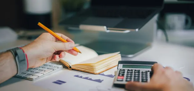 a person sitting at a desk with a calculator and a notebook by Jakub Żerdzicki courtesy of Unsplash.